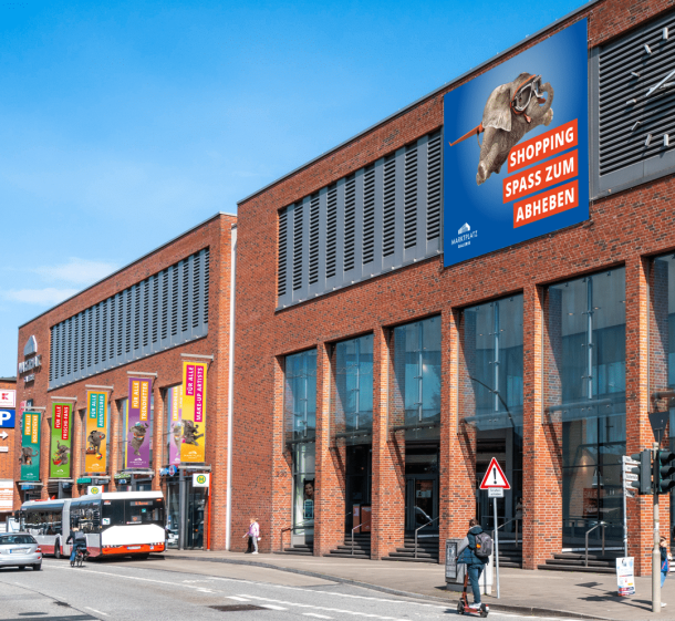 großes Außenbanner sowie Außenfahnen für die Marktplatz Galerie. Zu sehen ist das Center-Maskottchen in verschieden Positionen mit jeweils passender Headline